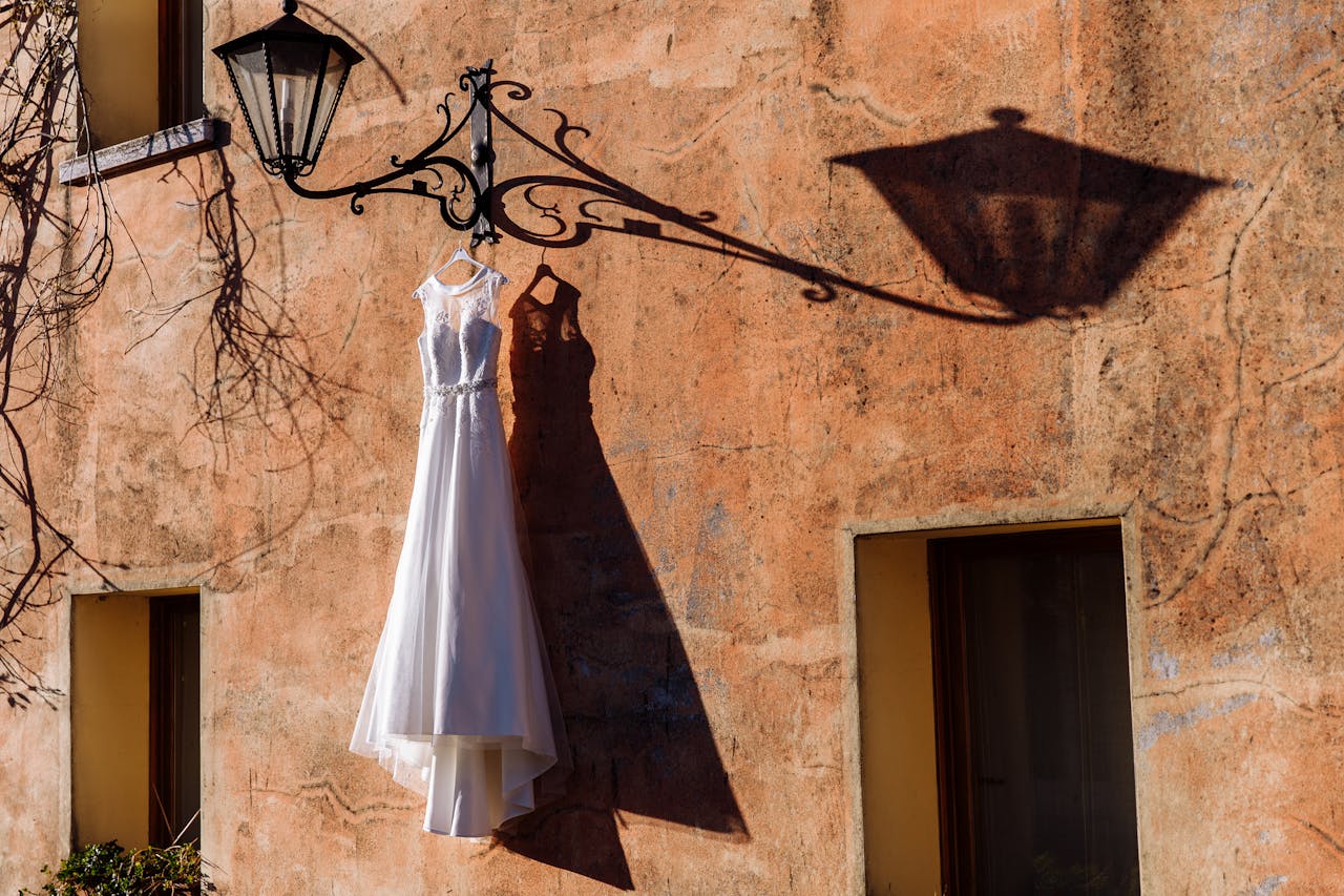 A wedding dress hangs from an ornate lamp in Rome, casting a dramatic shadow on a rustic wall.