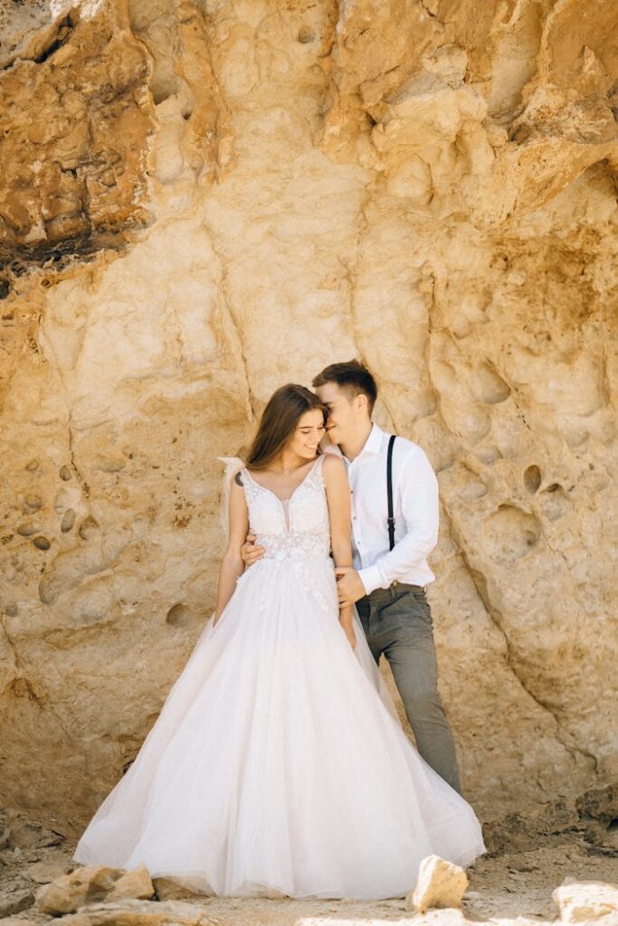 A beautiful couple in wedding attire sharing a tender moment against a rustic cliff backdrop.